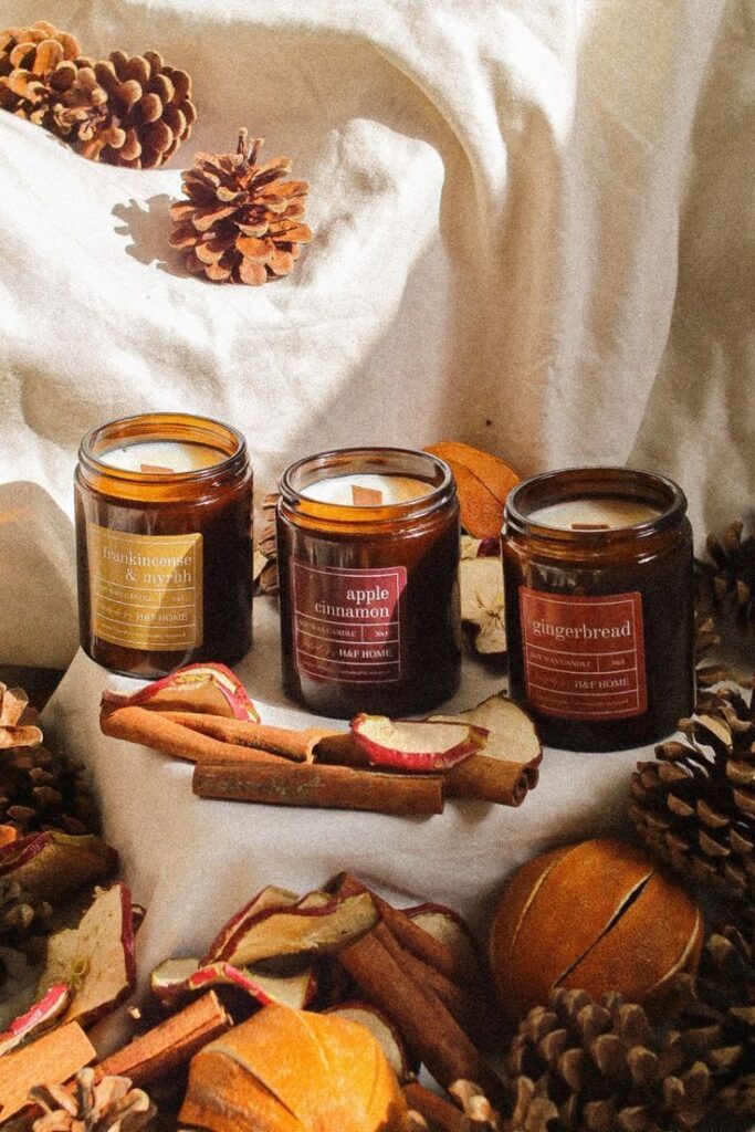 Cozy scented candles with cinnamon sticks and pine cones on white fabric, evoking a warm autumnal ambiance.