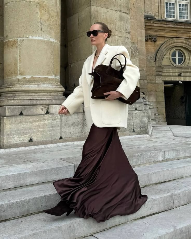 Fashionable woman in white blazer and flowing burgundy skirt walking down stone steps with a brown bag.