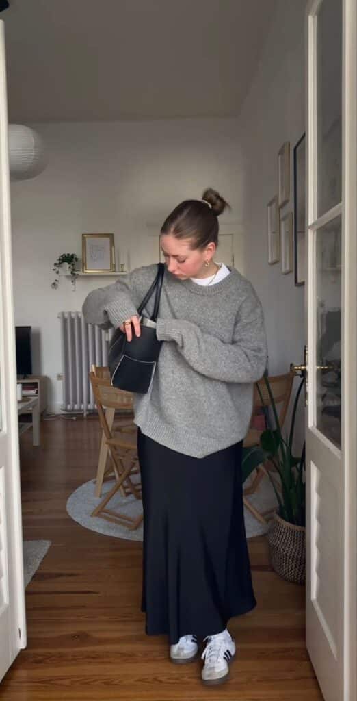 Woman in cozy outfit, gray sweater and black skirt, searches purse in stylish room with wooden floors.