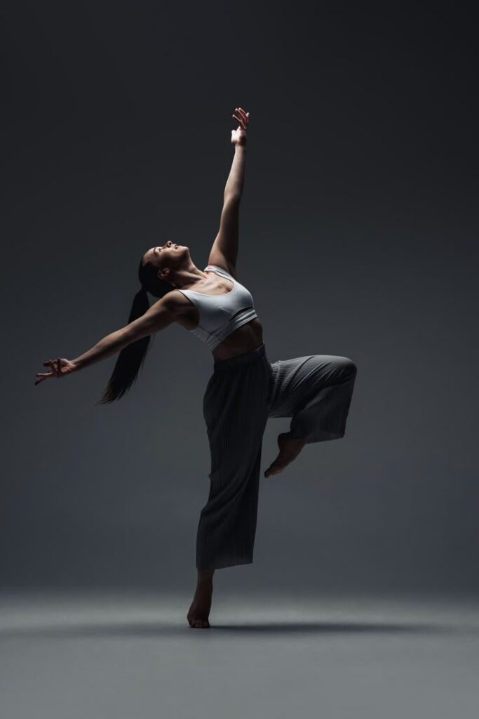 Dancer performing a graceful pose in a spotlight, wearing a white top and gray pants.