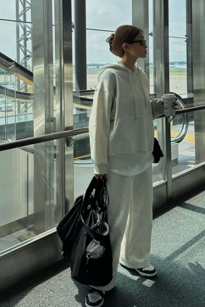 Person in a cozy airport outfit holding a coffee, carrying a black bag, standing by a window with a view of the runway.