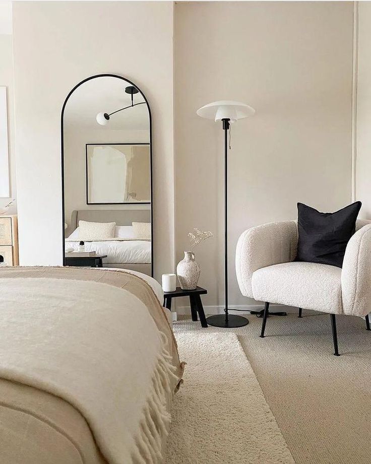 Cozy neutral bedroom with a modern armchair, floor lamp, and large mirror reflecting a neatly made bed. Minimalist decor. Minimalism and boho style