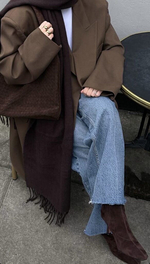 Stylish winter outfit with brown coat, maroon scarf, blue jeans, and boots, accessorized with rings and woven bag.