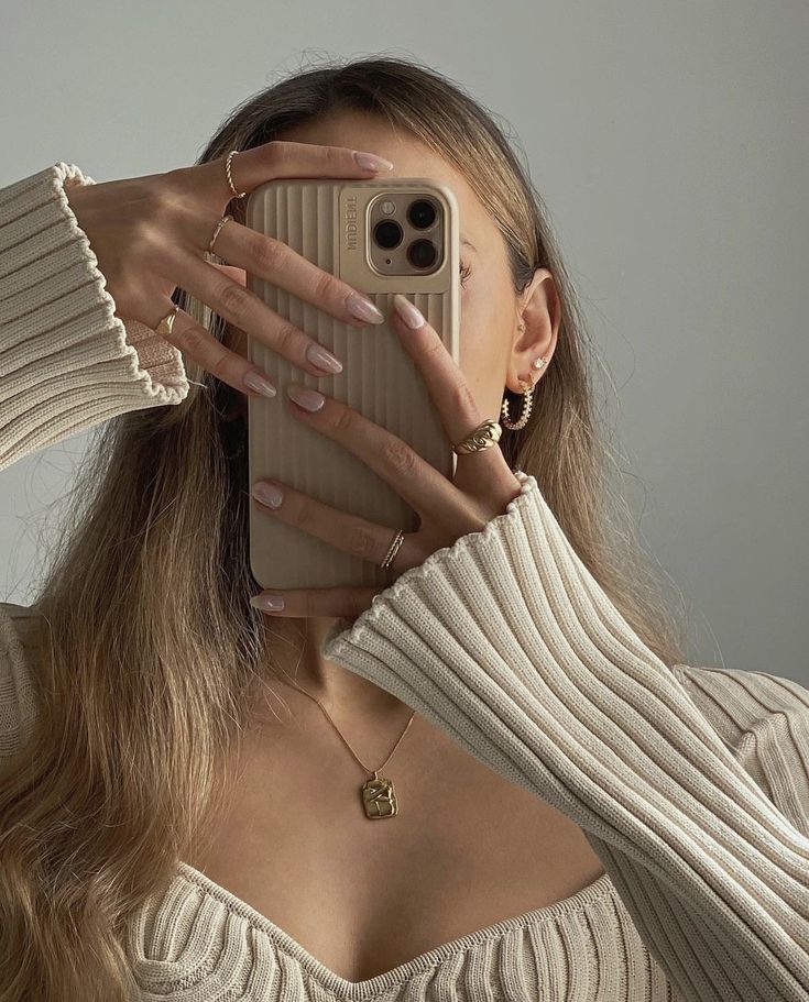 Person taking a selfie with a beige ribbed phone case, wearing gold jewelry and a knit top.