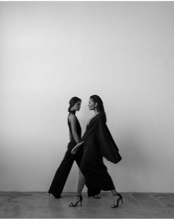 Two women walking in elegant black outfits, minimalist setting, monochrome fashion photography.