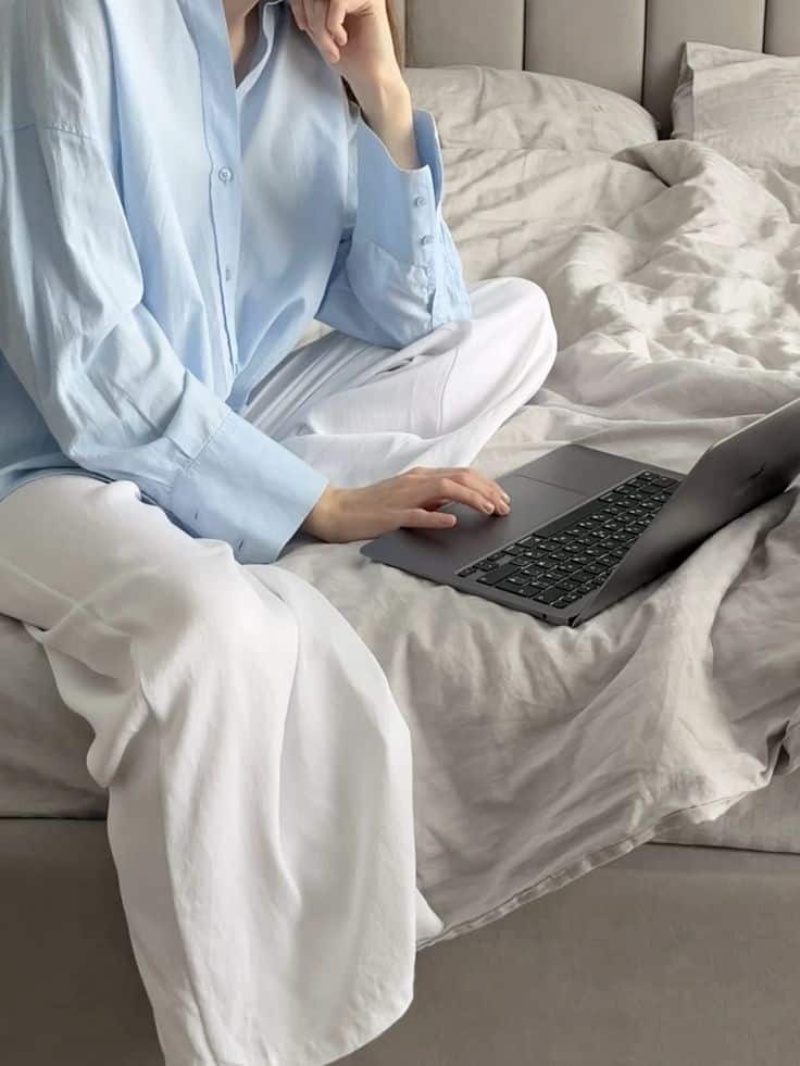 Person in casual attire working on a laptop from their bed, promoting remote work flexibility and comfort.
