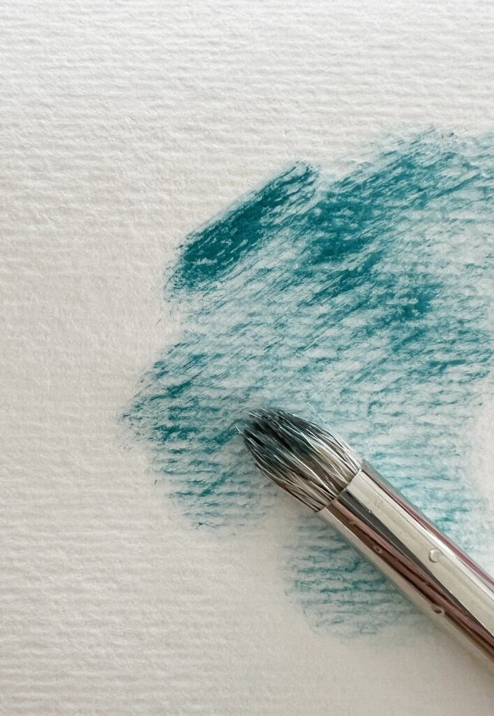 Paintbrush with teal strokes on white textured paper, showcasing watercolor technique.