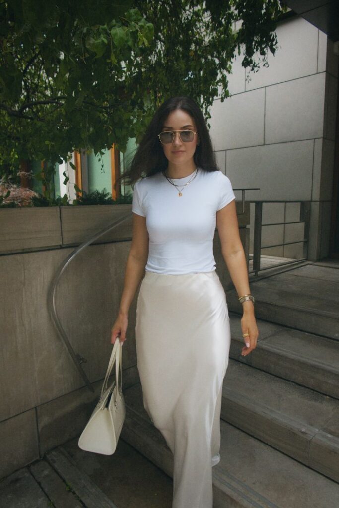 Woman in a white top and satin skirt holding a bag, walking down stairs in a garden setting.