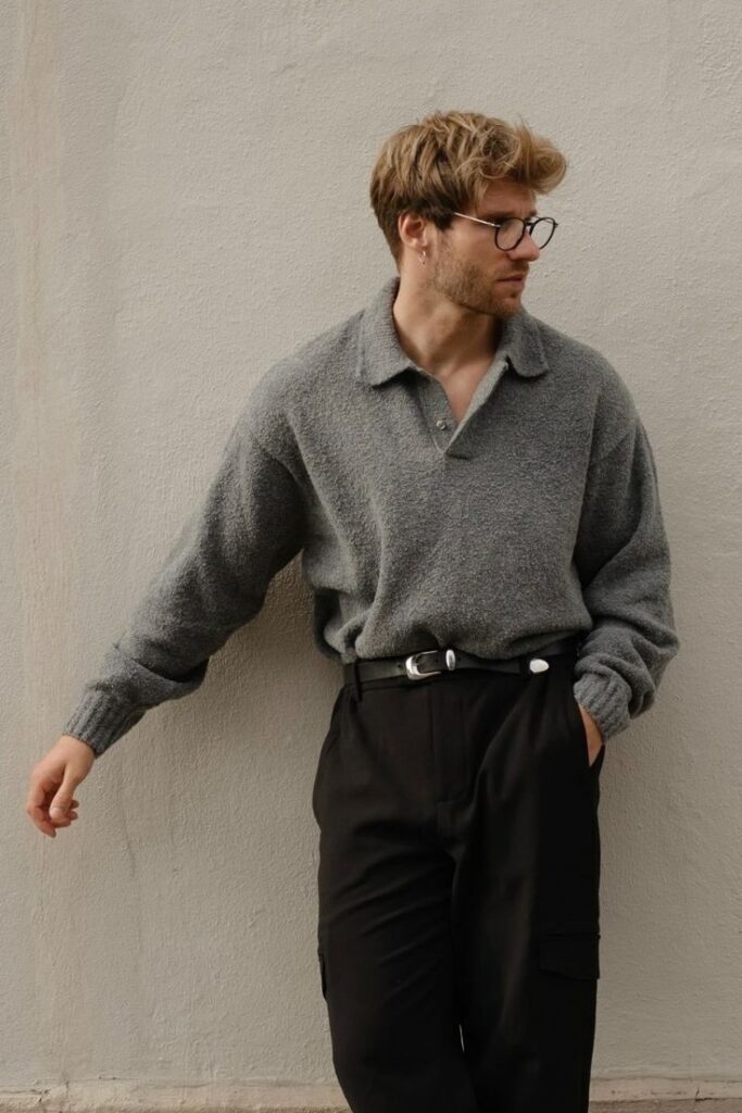 Man in grey sweater and black pants leans against a wall, wearing glasses, styled in casual fashion.