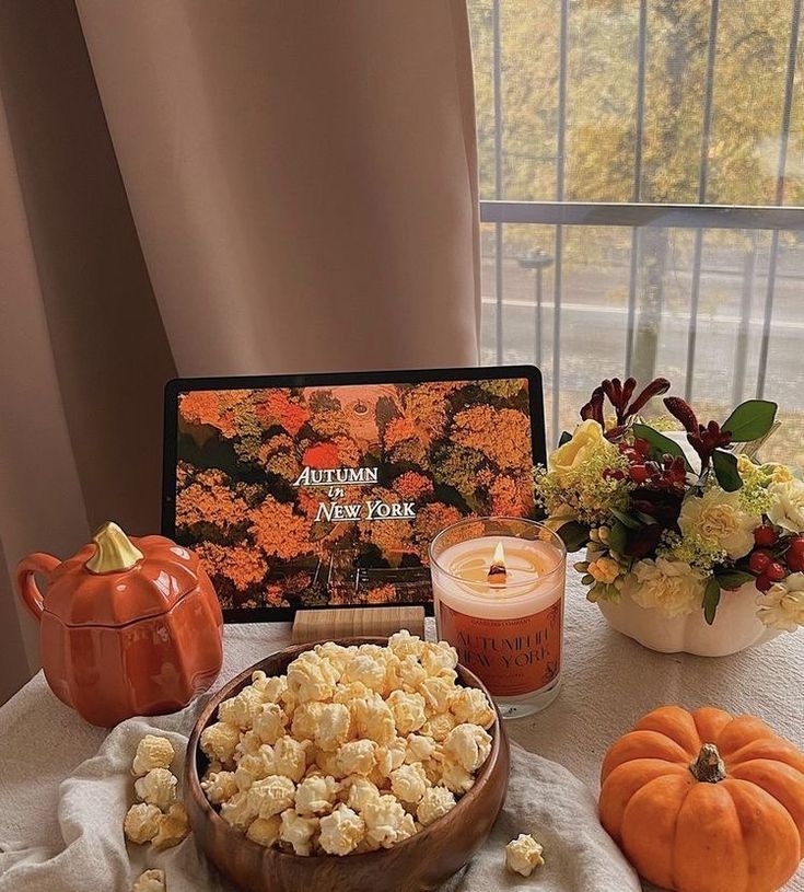 Cozy autumn setup with popcorn, pumpkin decor, candle, flowers, and Autumn in New York movie on tablet.