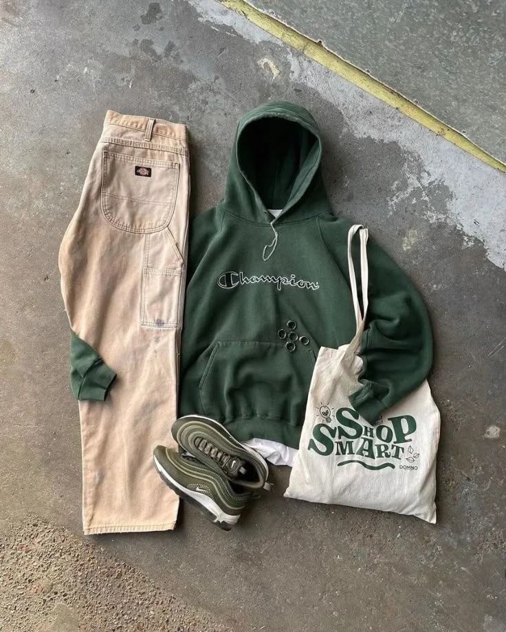 Casual outfit flatlay with green hoodie, beige pants, sneakers, and a tote bag on concrete floor.