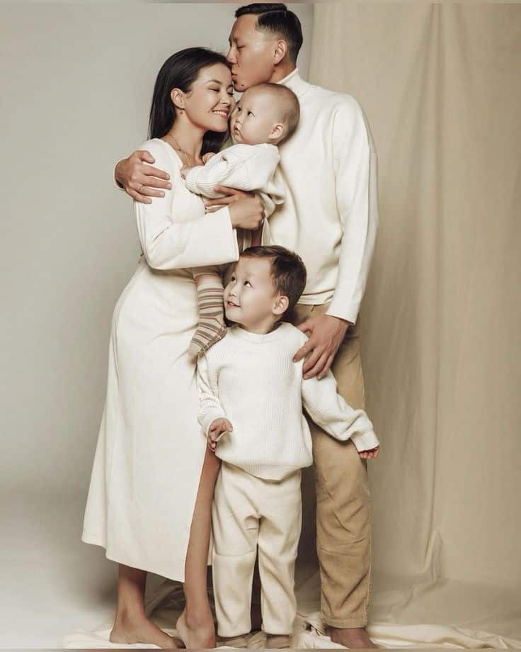Happy family of four in cozy cream outfits sharing a loving embrace in a warm, neutral setting.