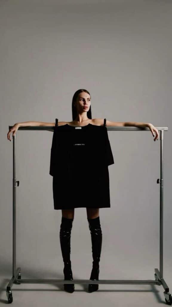 Model wearing black boots with arms on garment rack, displaying black oversized hoodie. Minimalist fashion.
