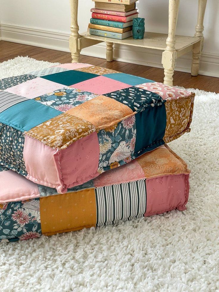 Two stacked colorful patchwork floor cushions on a white rug, next to a small table with books and a teal ornament.