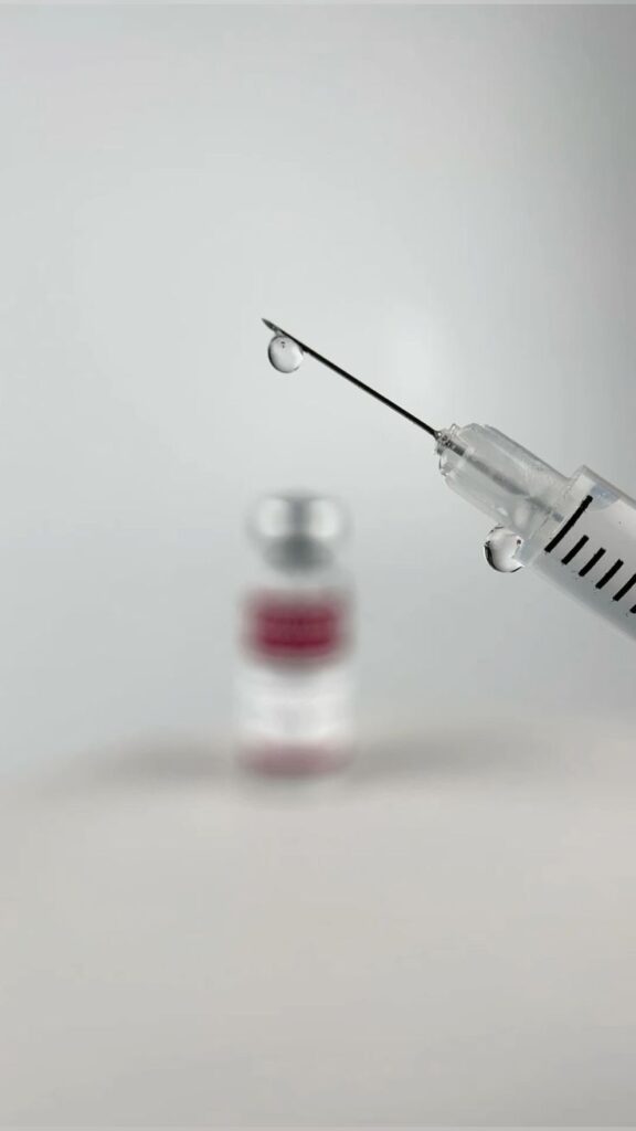 _ 3 Close-up of a syringe needle with a droplet, blurred vaccine vial in the background, on a neutral background. | Sky Rye Design Close-up of a syringe needle with a droplet, blurred vaccine vial in the background, on a neutral background.