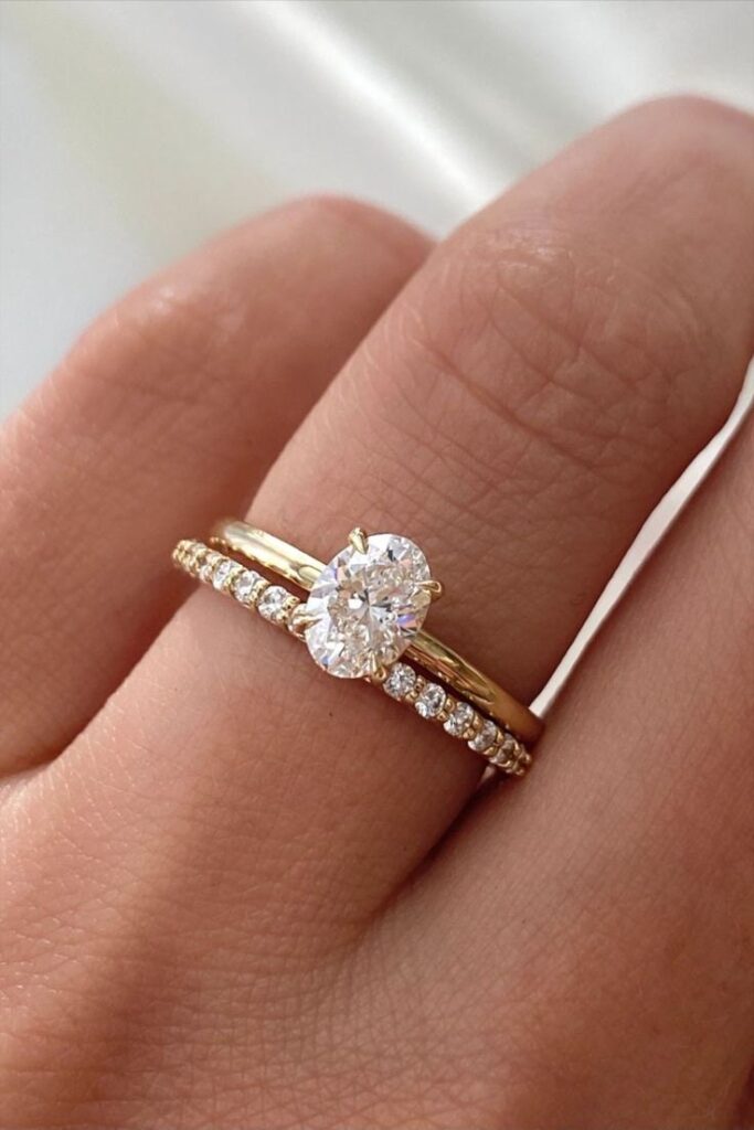 Elegant gold engagement ring with oval diamond, paired with a diamond-encrusted wedding band on a hand.