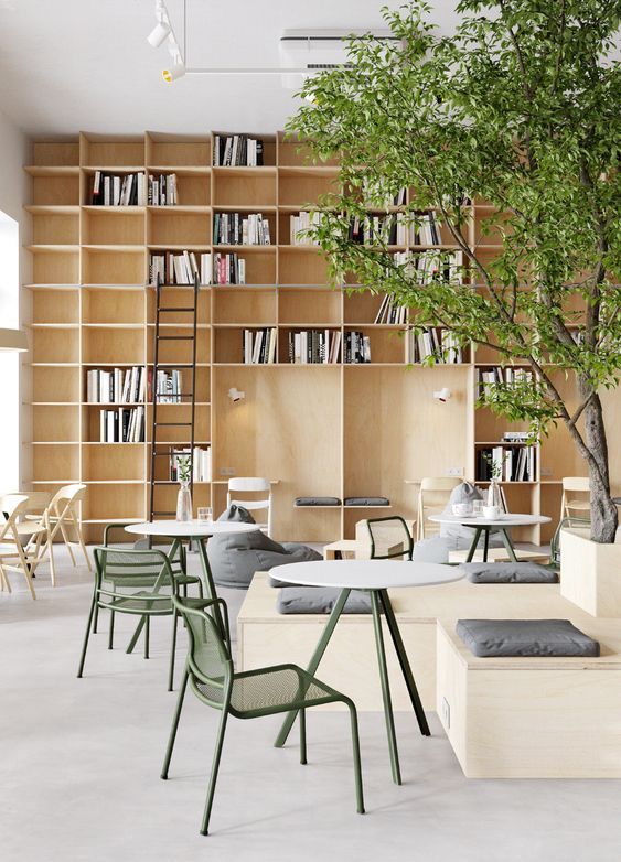 Modern library cafe with wooden bookshelves, green chairs, and indoor tree, creating a cozy reading space.