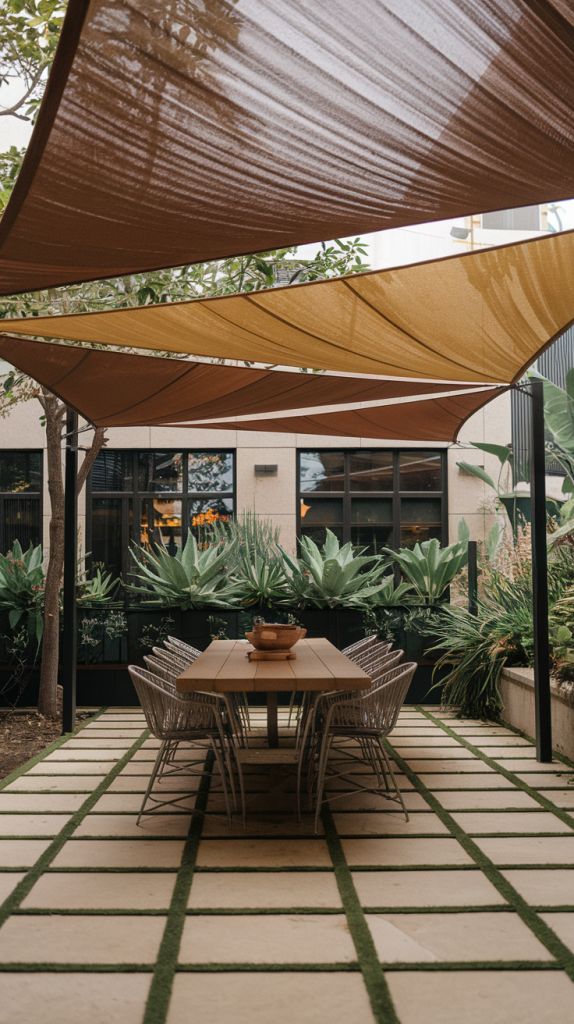 Outdoor patio dining area with modern table, chairs, shade sails, and lush greenery.