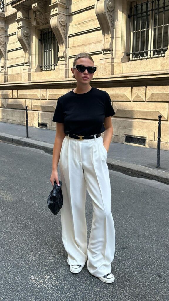 Chic woman in black tee and white pants with sunglasses, holding a clutch on a stylish city street. Urban fashion.