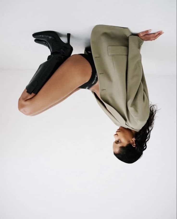 Model in a fashion pose wearing a beige jacket, black boots, and underwear against a white background.