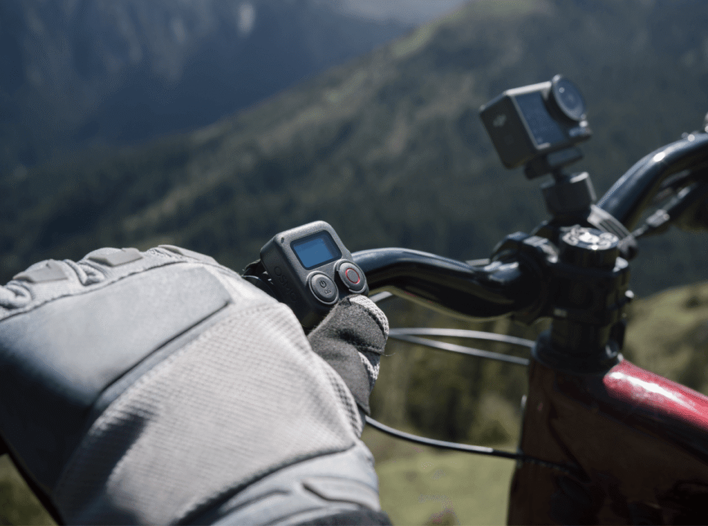 Gloved hand holding action camera on bike handlebar, capturing mountain scenery in the background.