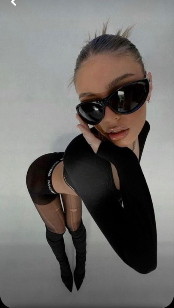 Fashionable woman in black outfit, sunglasses posing confidently against a minimalist background.