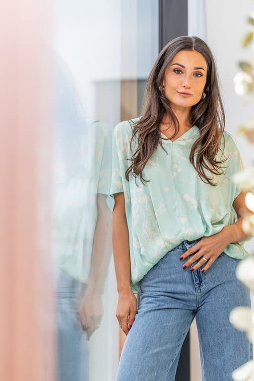 Woman in a light green blouse and jeans standing by a window, exuding casual elegance and confidence.