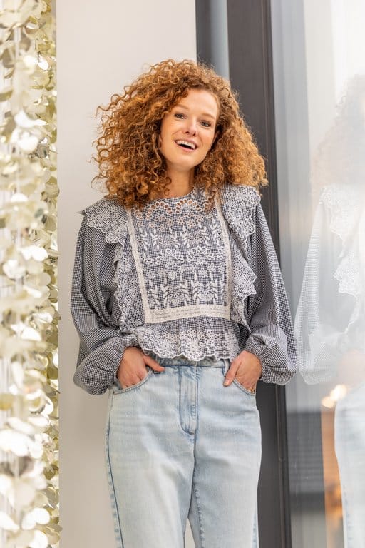 Smiling woman with curly hair wearing a lace top and jeans, standing by a window in natural light.