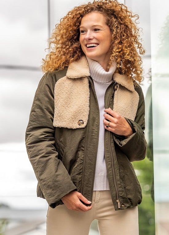 33649_5 Smiling woman in a cozy shearling-lined olive jacket, beige pants, and white turtleneck, enjoying a cloudy day outdoors. | Sky Rye Design Smiling woman in a cozy shearling-lined olive jacket, beige pants, and white turtleneck, enjoying a cloudy day outdoors.