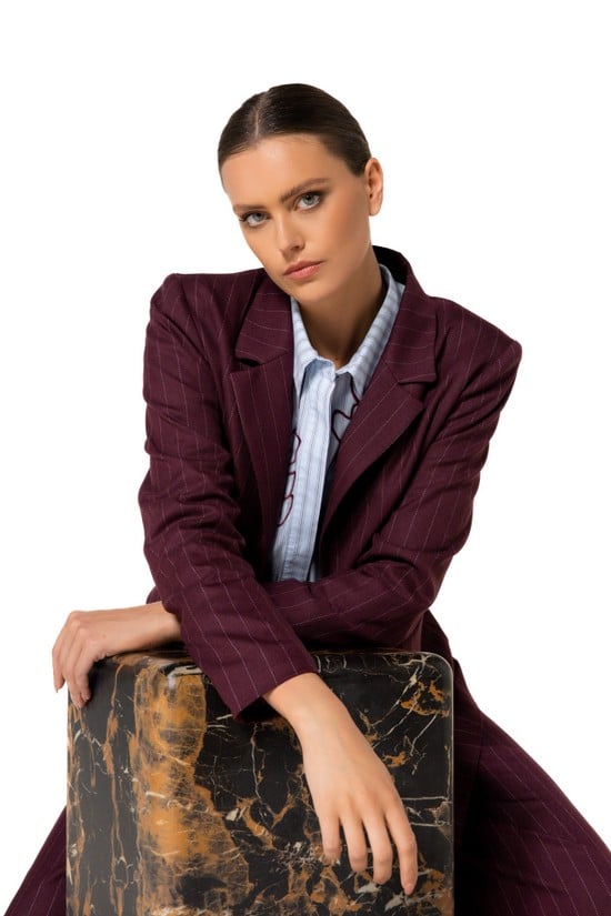 Professional woman in maroon suit poses confidently against a marble block on white background.