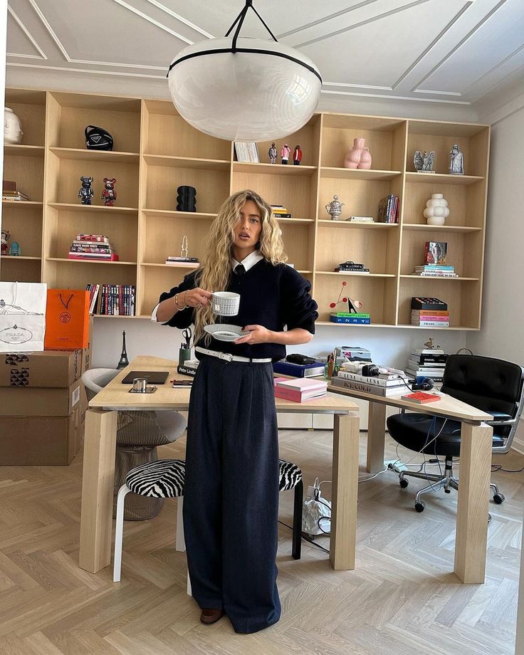 Person in a stylish office holding a cup, surrounded by books and decor on wooden shelves.