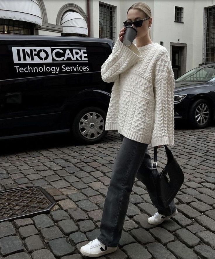 Fashionable woman in cozy sweater and jeans walking with coffee on cobblestone street, carrying a black handbag.