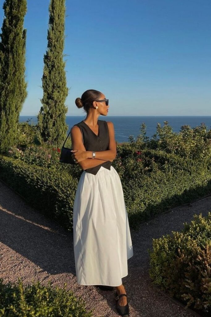 Woman in stylish black top and white skirt poses on a garden path by the sea under clear blue skies.