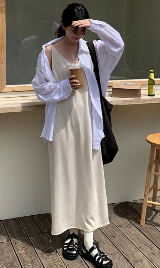 Woman in a white dress and cardigan holds iced coffee, standing outdoors, shielding her eyes from the sun.