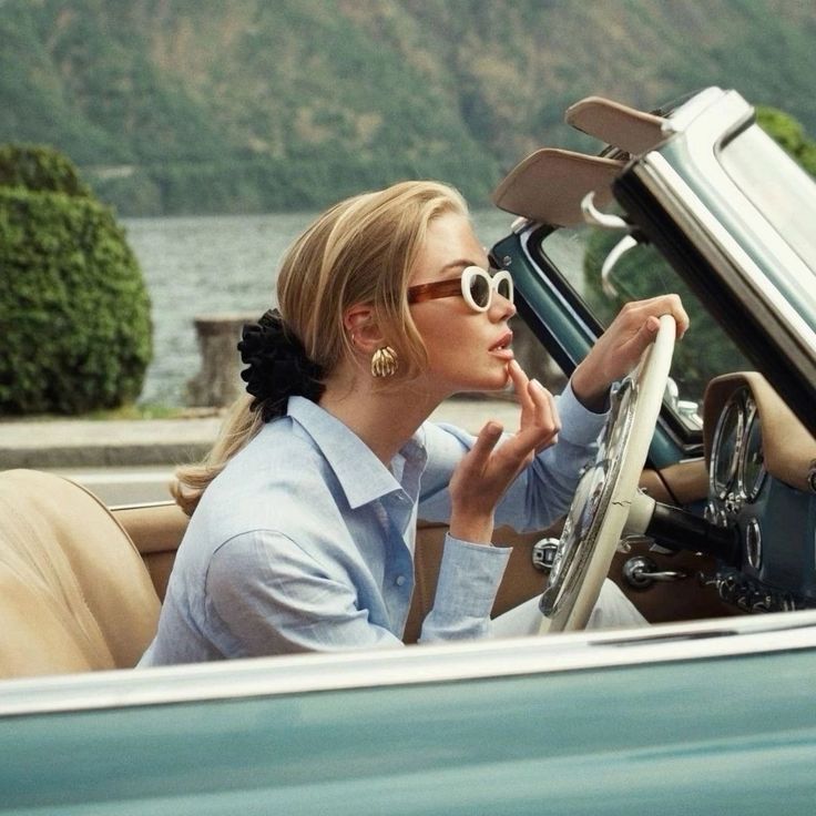 3eb020da-11e3-459d-955b-c702b050c5ad Woman in sunglasses applying lipstick in a vintage convertible car by a scenic lake. | Sky Rye Design Woman in sunglasses applying lipstick in a vintage convertible car by a scenic lake.