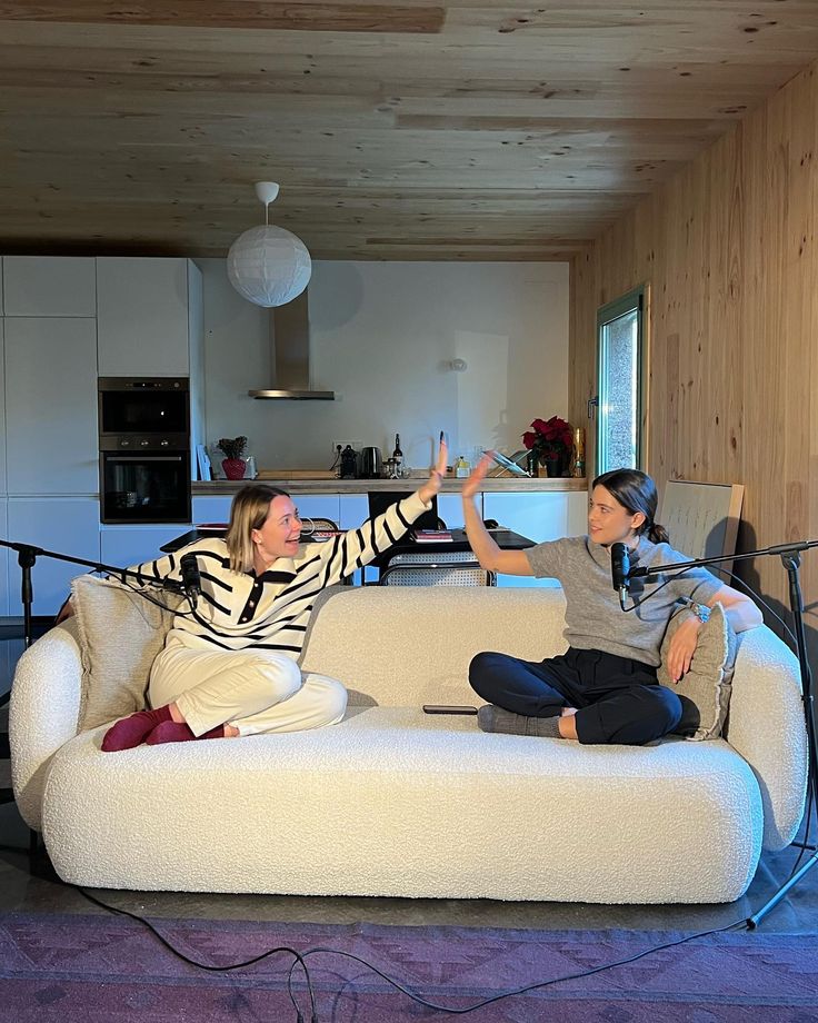 Two women high-five on a sofa during a podcast recording in a cozy, modern living room setting.