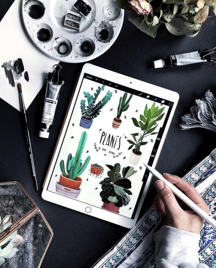 Digital illustration of houseplants on a tablet with art supplies and a hand holding a stylus.