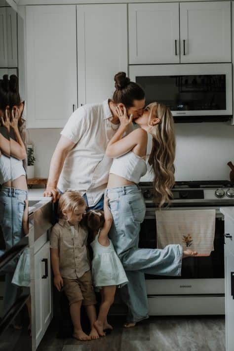 Happy family in kitchen: couple kissing, two young kids nearby; warm, modern home atmosphere.