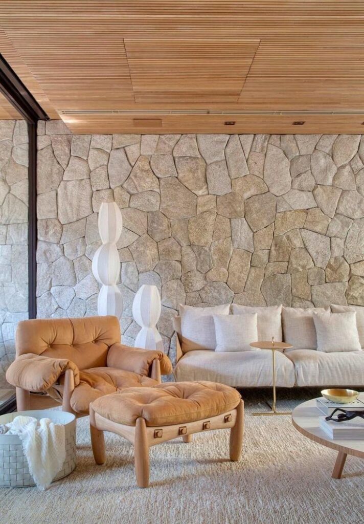Modern living room with stone wall, beige chair, sofa, round wooden coffee table, and stylish decor accents.