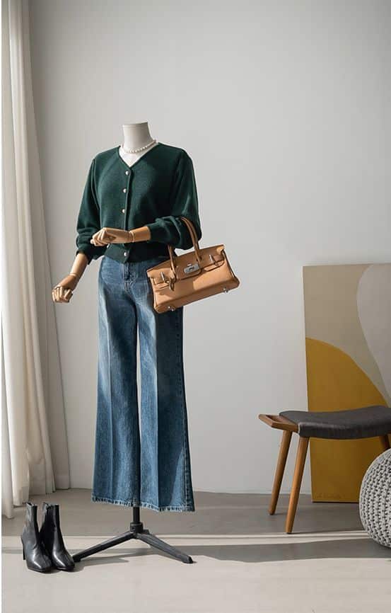 Mannequin in green cardigan, wide-leg jeans, and tan handbag, with black boots; light, modern room setting.