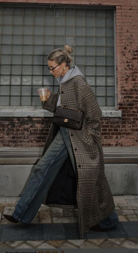 Stylish woman in plaid coat and jeans walking with iced coffee and designer handbag in urban setting.
