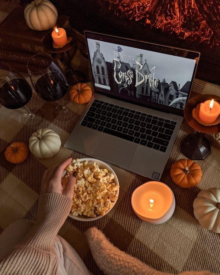 Cozy movie night setup with laptop, popcorn, candles, pumpkins, and wine, perfect for a Halloween-themed evening.