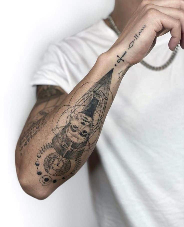 Arm with detailed geometric and portrait tattoo, wearing a white shirt and chain bracelet.