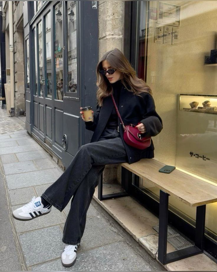 Stylish woman sitting on a bench, enjoying coffee, wearing sunglasses and casual chic outfit on a city street.