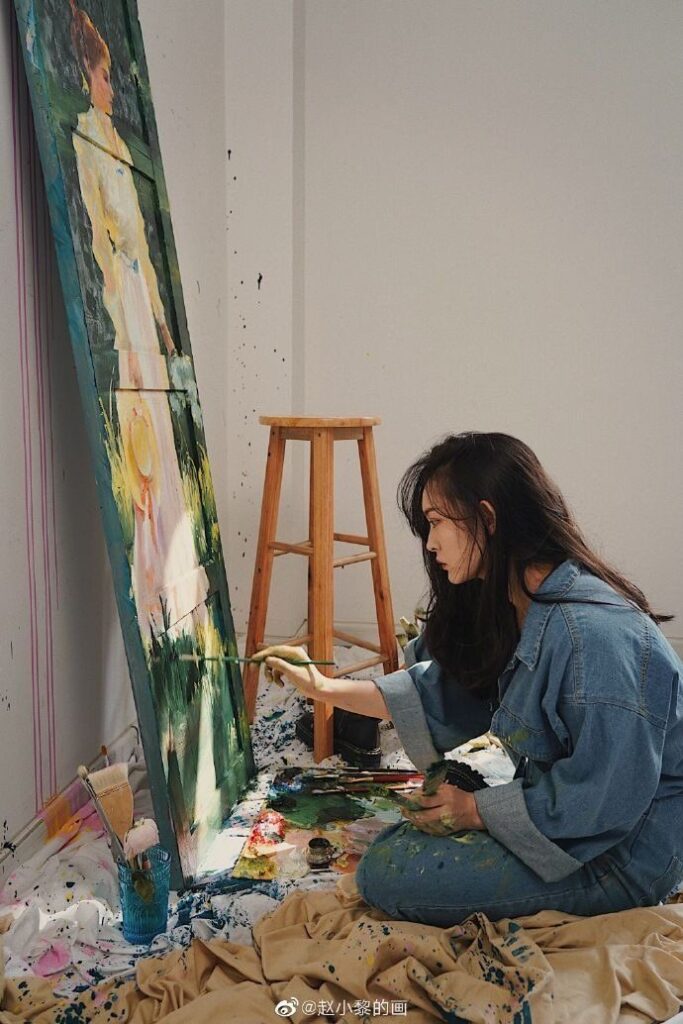 Artist painting a colorful canvas while sitting on a paint-splattered floor, wearing a blue denim outfit.