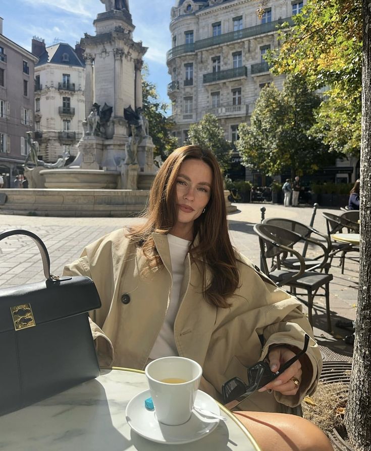 Woman enjoying coffee at a sunny outdoor café in Paris, stylish trench coat and city backdrop in view.