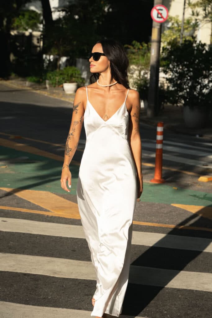 Woman in a white dress and sunglasses walking on a city street crosswalk.