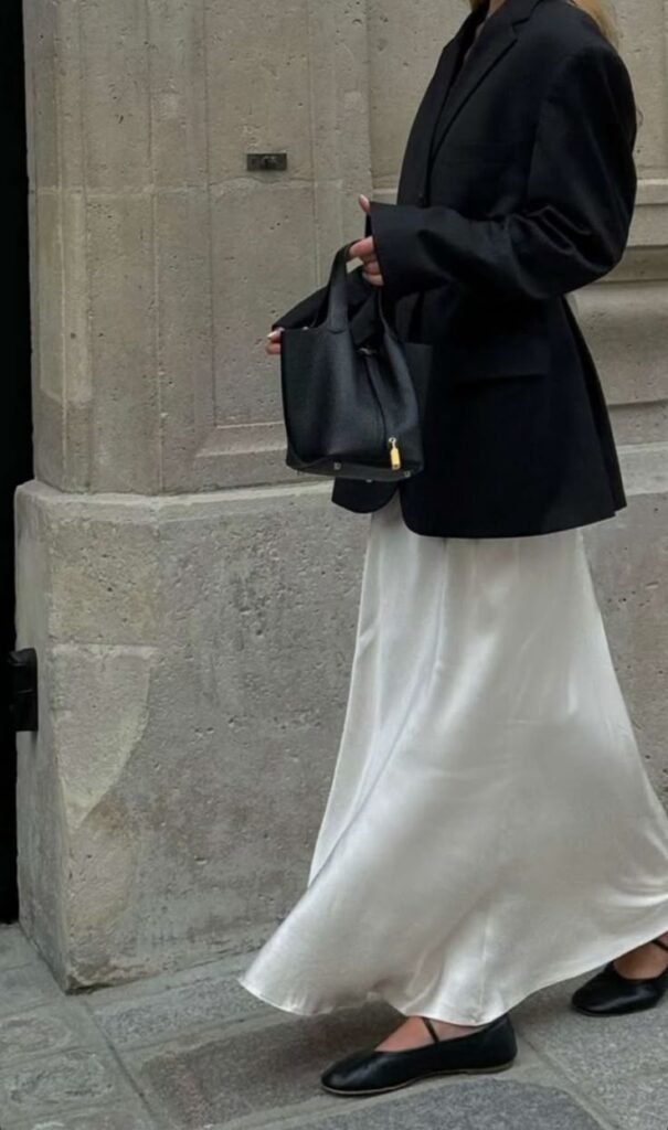 Woman in black blazer, white skirt, and flats holds a black handbag on a city street. Stylish and elegant fashion.