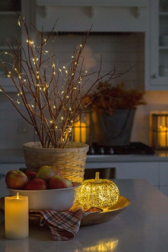 Cozy fall kitchen decor with glowing pumpkin, apples, candle, and fairy lights in a rustic setting.