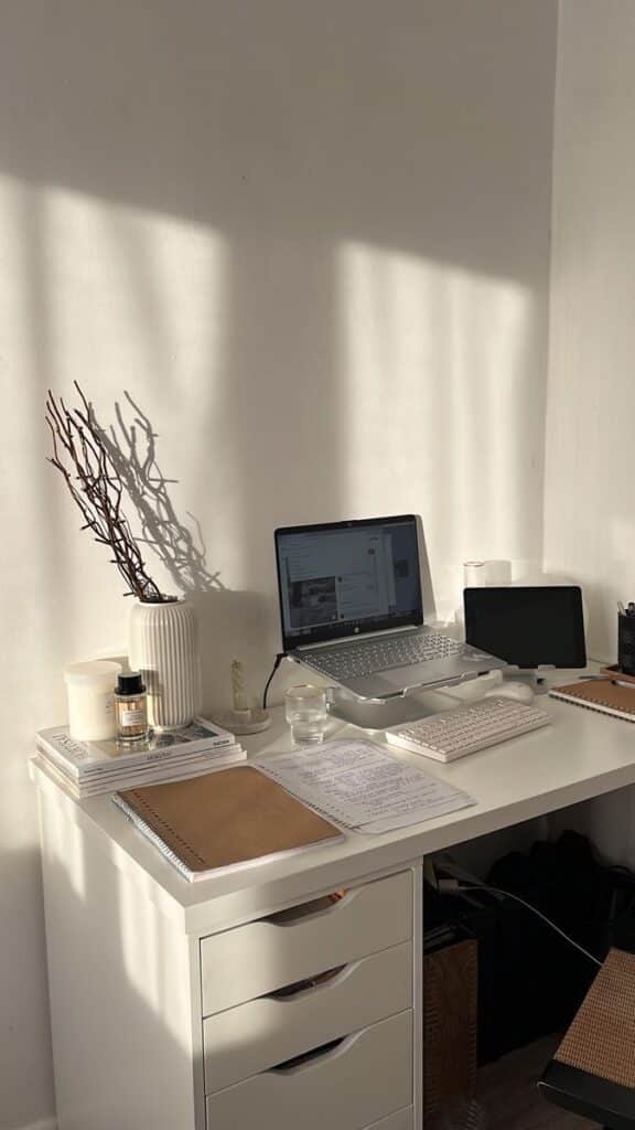 Minimalist home office desk setup with laptop, tablet, notebooks, and decor in natural lighting.
